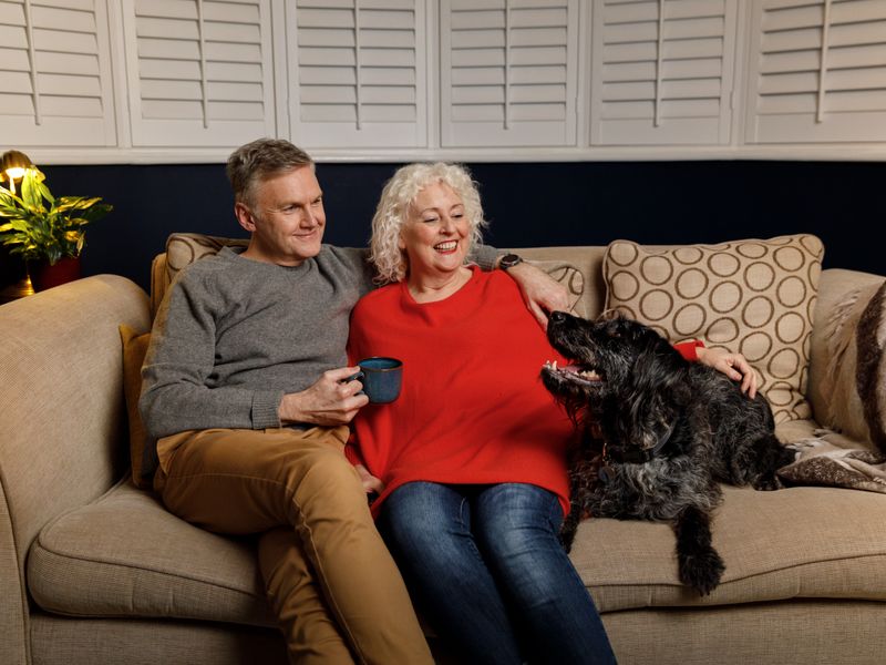 Man and woman sitting on a sofa together hugging, with a black long-haired dog.