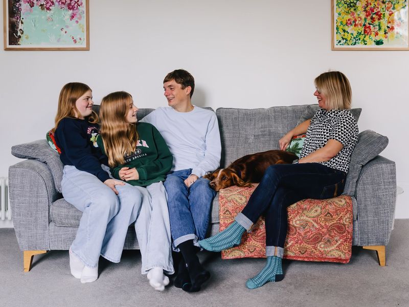 Family of four with mum, dad and two girls sitting and smiling at each other on a grey sofa with a orange spaniel type dog.