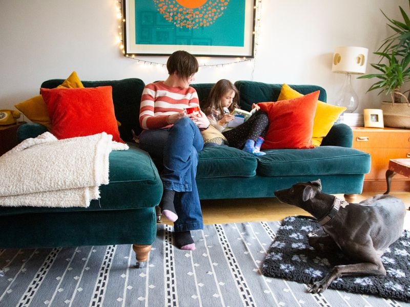 Adult female sitting on a teal sofa with orange and yellow cushions. She sits with a child who is reading and grey greyhound on the floor on a grey blanket.