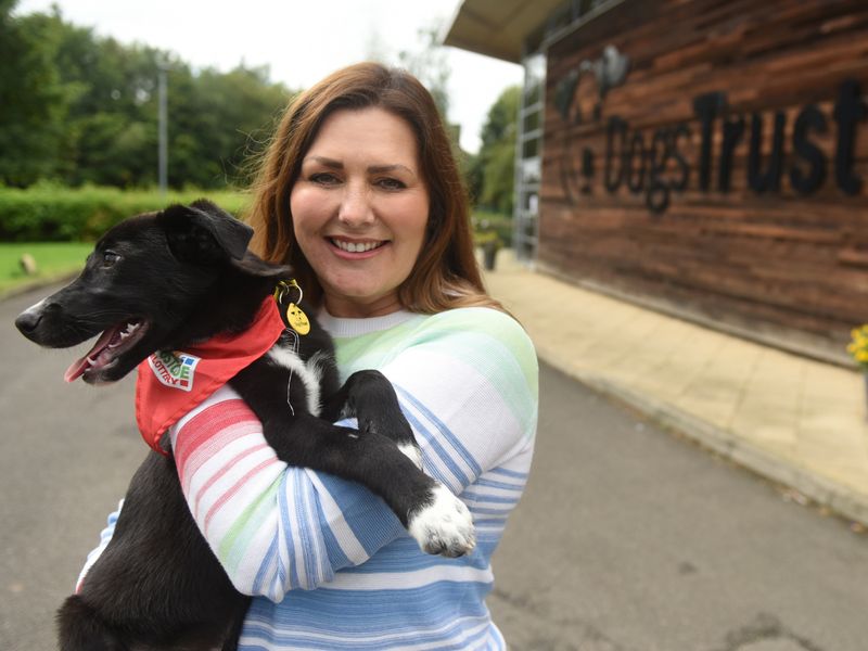 Photo of a lady holding a black crossbreed dog wearing a Peoples postcode lottery red bandana, outside a Dogs Trust centre.