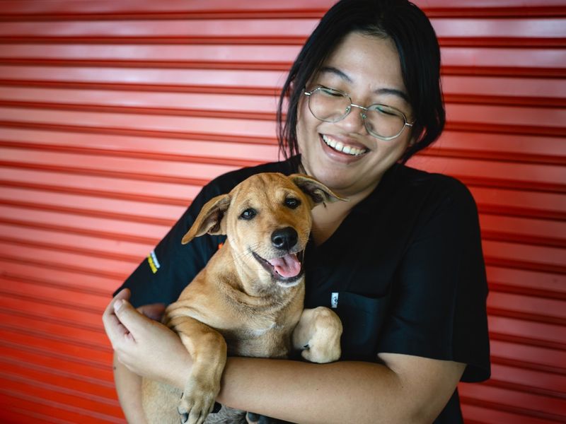 Woman holding and looking down at a tan puppy who's smiling with tongue out looking at the camera.