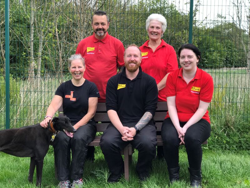 Dogs Trust West Calder volunteers and Volunteer Coordinator sitting with a black greyhound