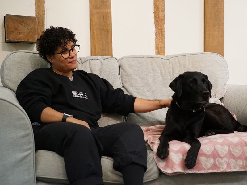 A lady with a hand on Coco a black Labrador laying on a pink blanket sitting on a sofa