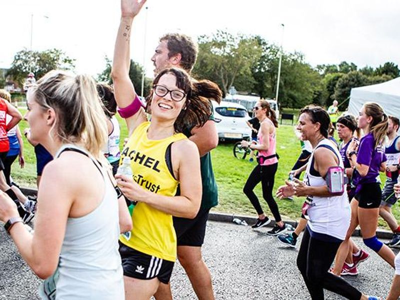 A woman running in a yellow dogs trust vest and putting her thumb up in the air and smiling at the camera, running with a sea of runners at the Edinburgh Marathon