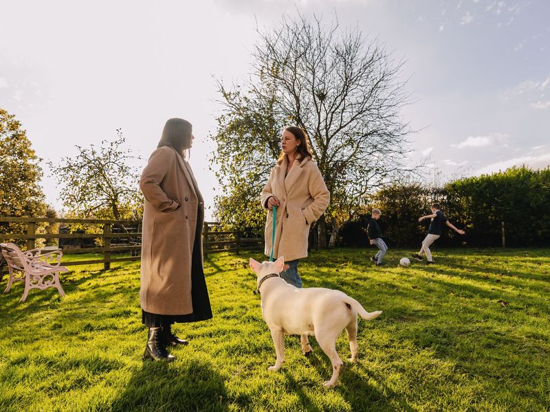 A white bull terrier standing next to two ladies chatting in a park, whilst two boys play football in the background