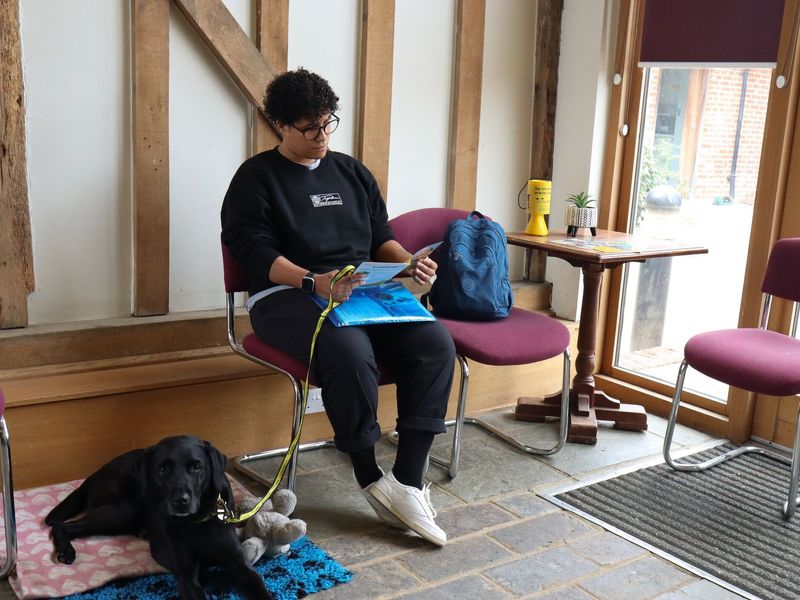 Coco a black labrador, sitting on the floor of a vet clinic waiting room beside her owner who is looking at papers